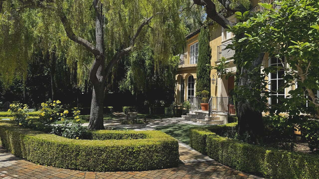 Clipped boxwood parterre and yellow roses curving through a brick motor court beneath mature willows, in front of a two-storey villa.